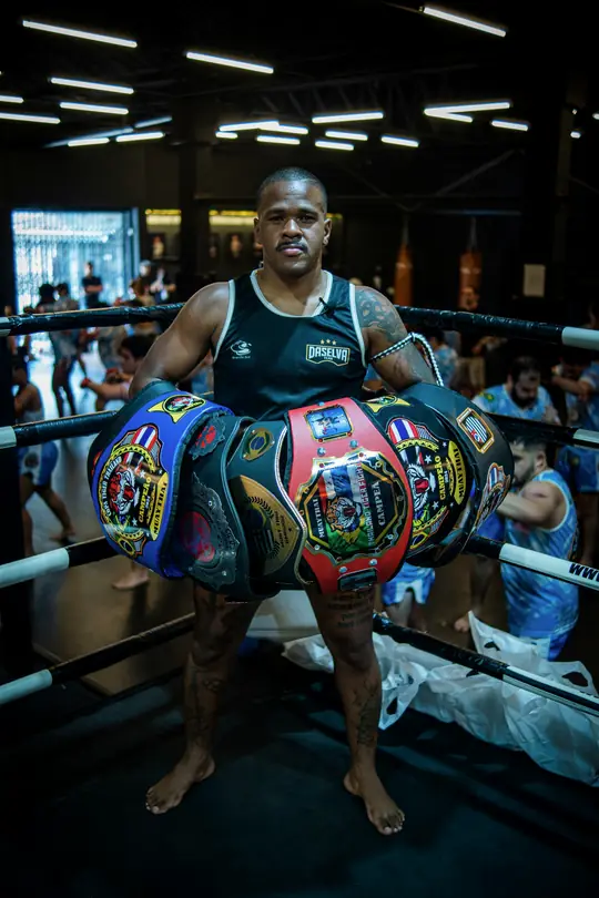 Professor Rubens no ringue segurando os cinturões conquistados pelos atletas da DaSelva Team, com alunos treinando ao fundo